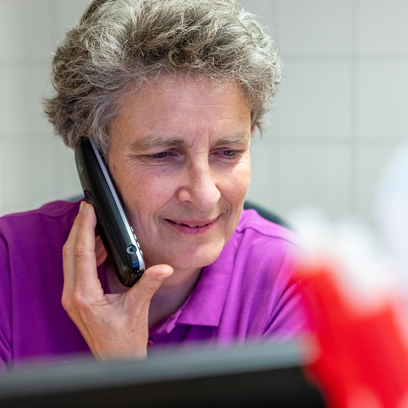 Eine Person mit kurzen, lockigen grauen Haaren und einem lila Hemd lächelt, während sie in einem Raum vor einem Computerbildschirm sitzt und in ein schnurloses Telefon spricht.