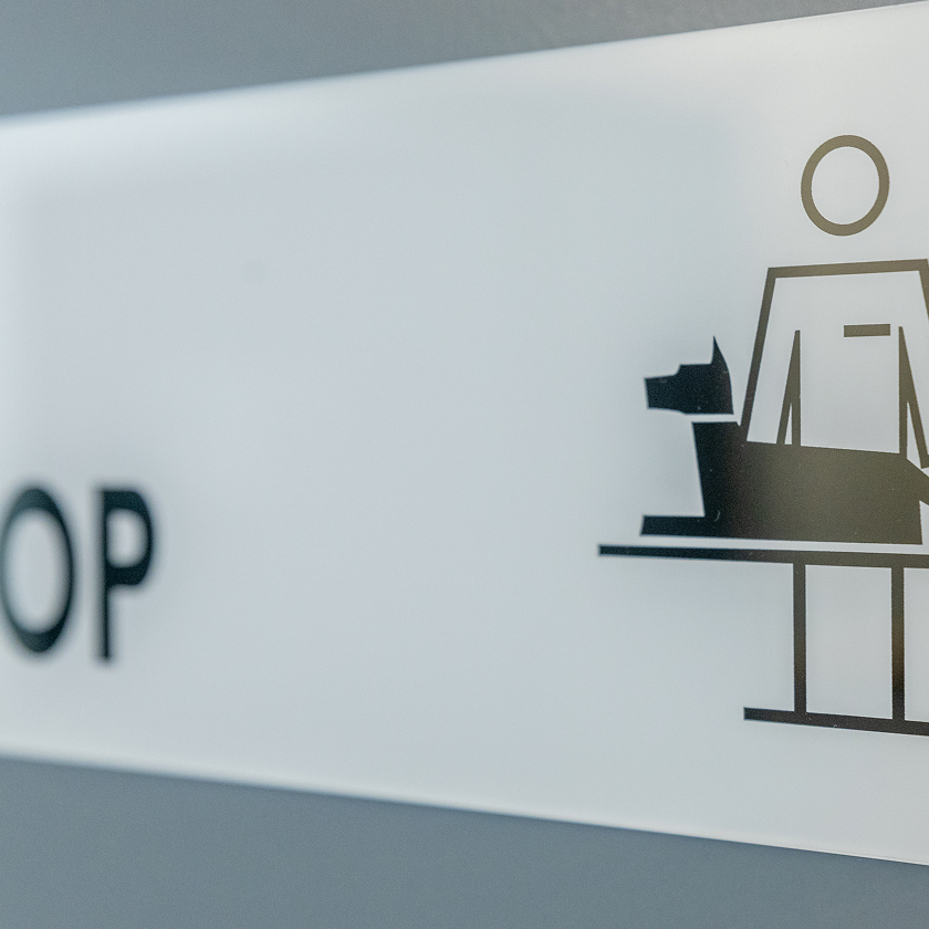 Ein Schild mit einer einfachen Illustration einer Person, die neben einem Hund auf einem Untersuchungstisch steht, weist auf eine Tierklinik oder einen Bereich für Haustiere hin.