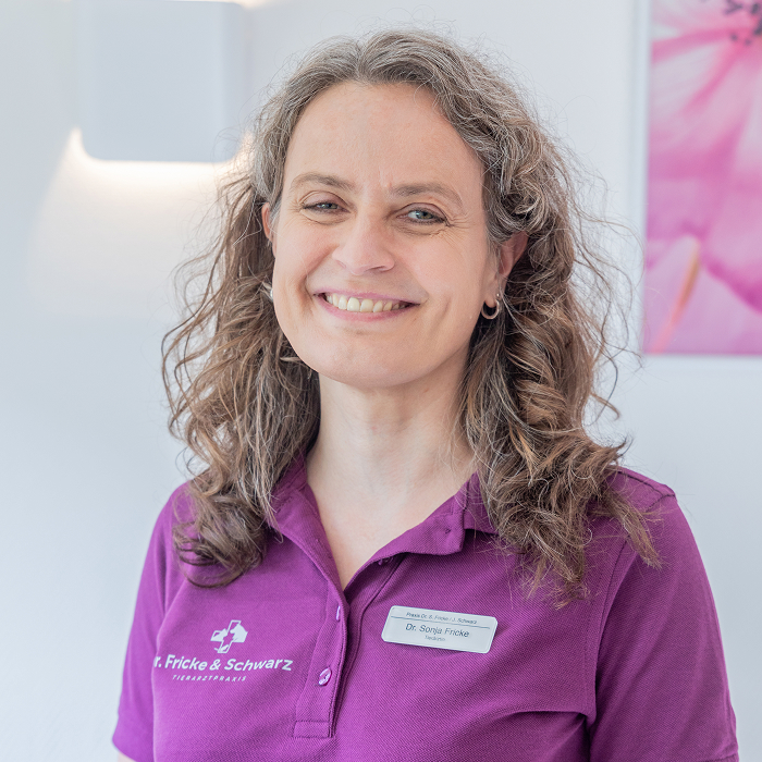 Eine Frau mit lockigem Haar lächelt in die Kamera. Sie trägt ein lilafarbenes Poloshirt mit einem Namensschild, auf dem "Dr. Sonja Fricke" steht, in einer hellen, professionellen Umgebung.