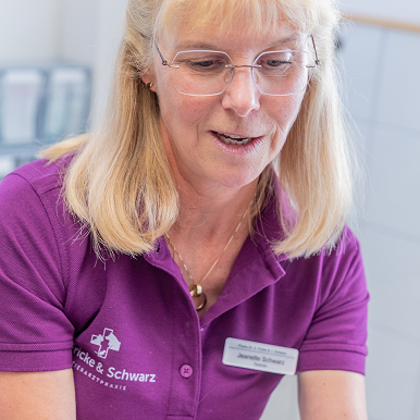 Eine Frau mit blondem Haar und Brille, die ein lila Hemd mit einem Namensschild trägt, scheint in einem medizinischen oder klinischen Umfeld zu arbeiten. Das Hemd trägt ein Logo und die Aufschrift "Fricke & Schwarz".