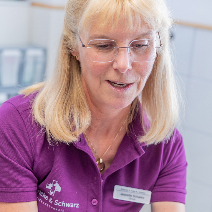 Eine Frau mit langen blonden Haaren und Brille, die ein lilafarbenes Hemd mit einem Logo und einem Namensschild trägt, blickt nach unten, möglicherweise in ein Gespräch vertieft oder bei der Arbeit in einer Klinik oder einem Büro.