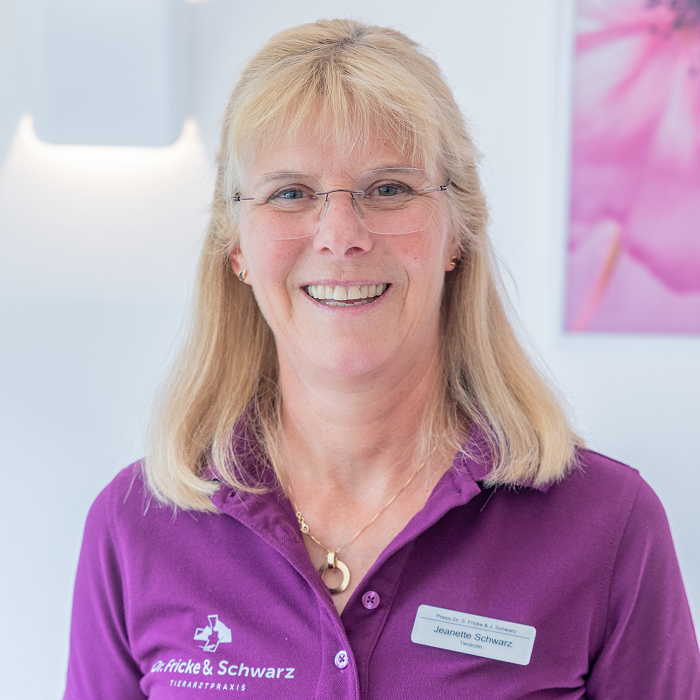 Eine lächelnde Frau mit blondem Haar, Brille und lila Kragenhemd mit Namensschild steht in einem hellen, modernen Raum mit hellen Wänden und einem rosa Kunstwerk im Hintergrund.