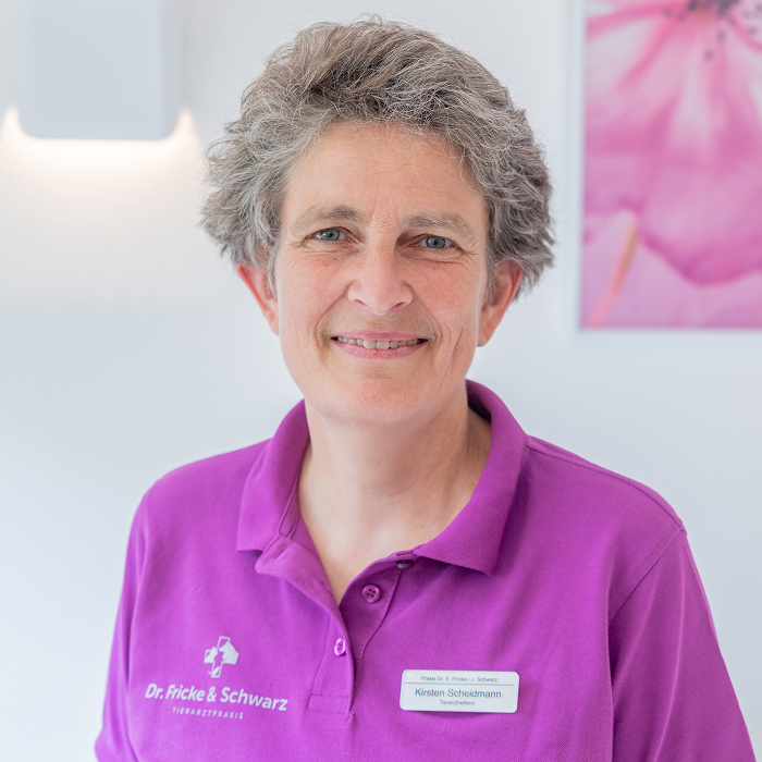 Eine lächelnde Frau mit kurzen grauen Haaren, die ein lila Polohemd und ein Namensschild trägt, steht in einem hellen Raum mit einem verschwommenen rosa Blumenbild an der Wand hinter ihr.