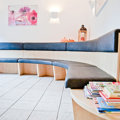 Ein Wartezimmer mit geschwungenen Bänken, einem gefliesten Boden, einem Stapel bunter Zeitschriften auf einem runden Tisch, Wandkunst mit rosa Blumen und sanfter weißer Beleuchtung.