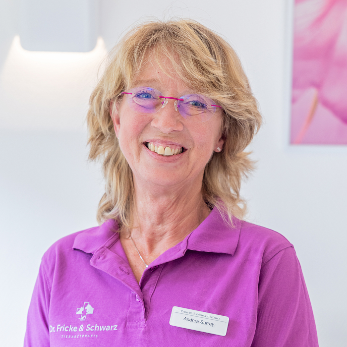 Eine lächelnde Frau mit blondem Haar und Brille trägt ein lila Poloshirt mit dem Logo "Dr. Fricke & Schwarz" und ein Namensschild mit der Aufschrift "Andrea Saurer". Sie steht in einem hellen Raum mit hellen Wänden.