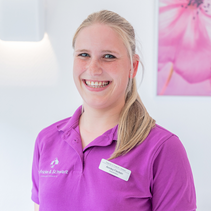 Eine Frau mit langen blonden Haaren in einem Pferdeschwanz lächelt, sie trägt ein lila Poloshirt und ein Namensschild. Sie steht in einem hellen Raum mit einem geblümten Bild an der Wand hinter ihr.