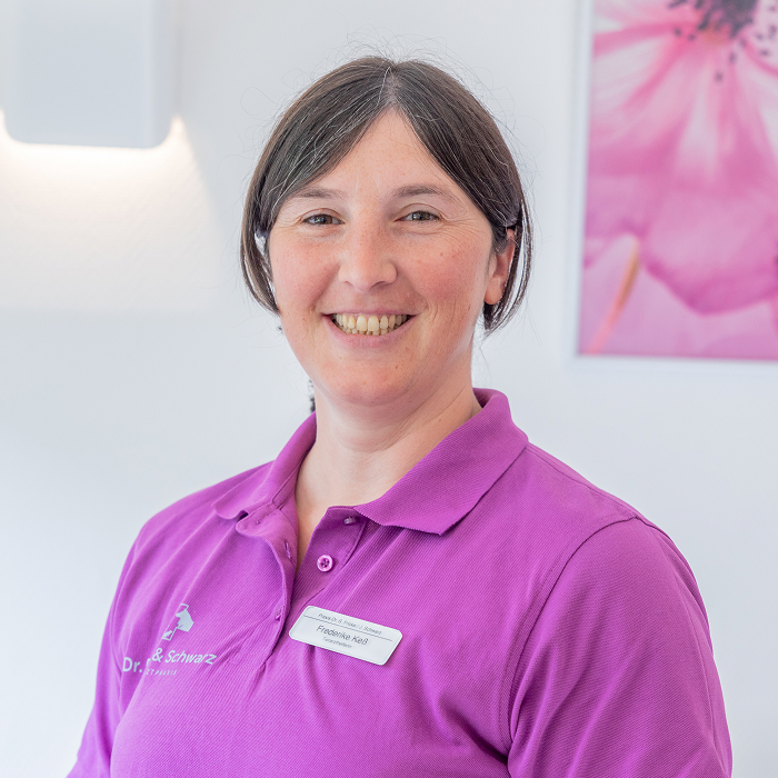 Eine Frau mit glattem braunem Haar und einem freundlichen Lächeln trägt ein lilafarbenes Hemd mit Kragen und ein Namensschild und steht in einem Raum neben einem verschwommenen rosa Blumenbild an der Wand.