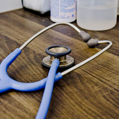 Ein blaues Stethoskop liegt auf einer hölzernen Oberfläche, im Hintergrund sind medizinische Geräte und Flaschen zu sehen.